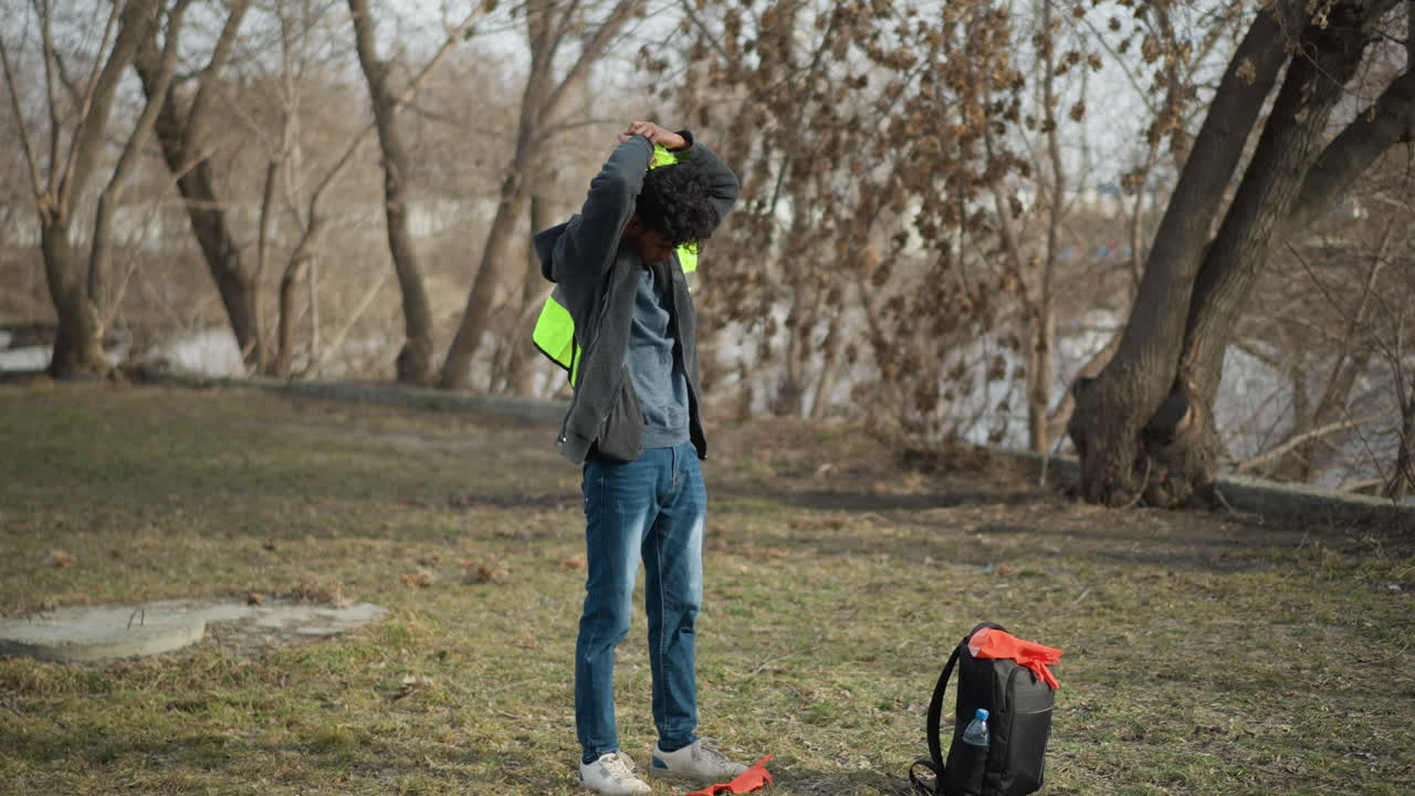 Man outdoors putting on bright orange protective gloves while holding neon green safety vest, preparing for outdoor cleanup activity in park with backpack nearby and bare trees in background
