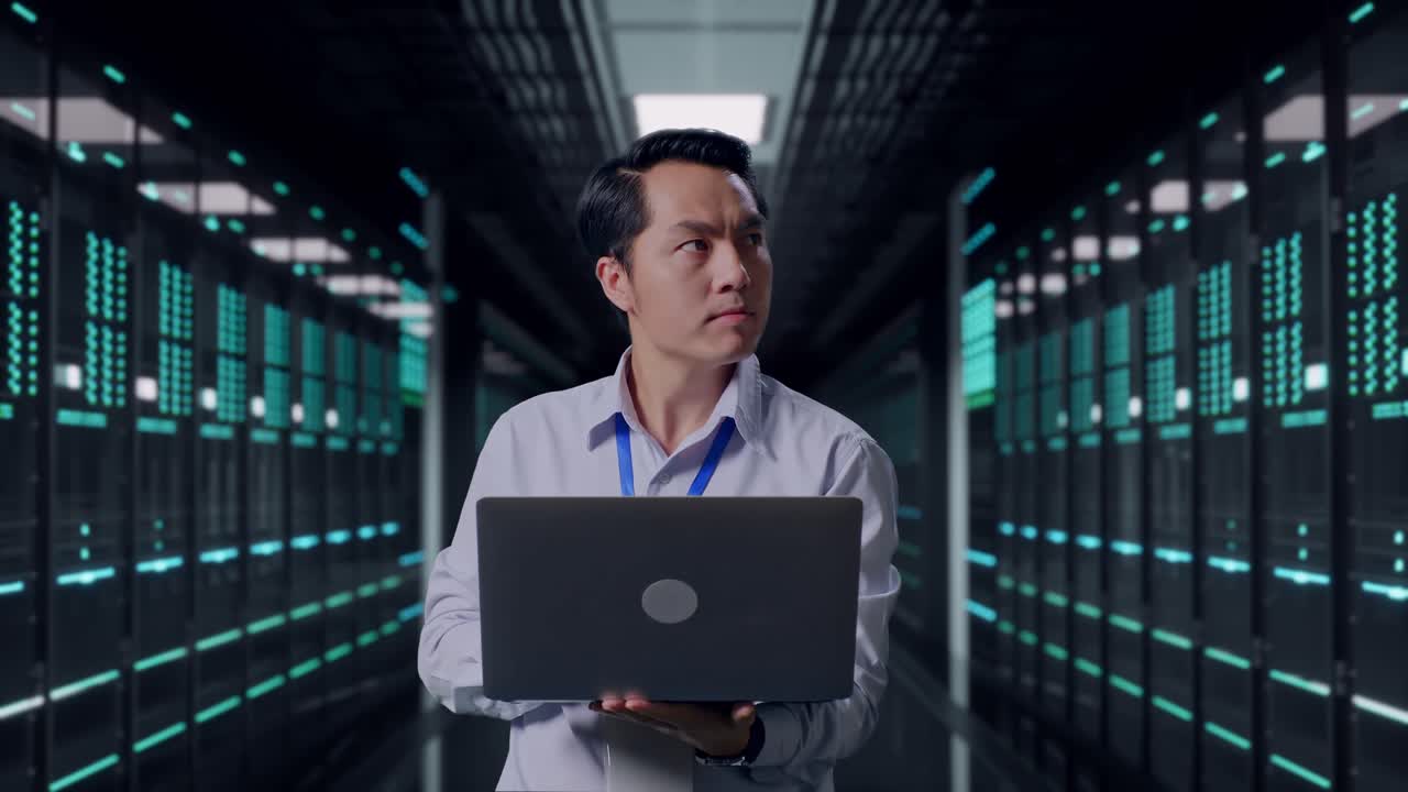 Asiatisk mandlig professionel arbejder, der står med sin bærbare computer i et datacenter. Han observerer ved at se sig omkring, før han koncentrerer sig om sin bærbare computer.