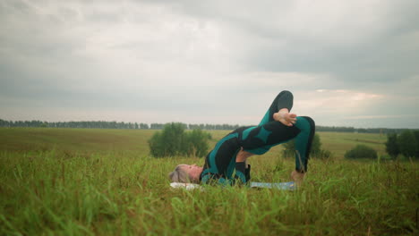 advanced woman in green black suit lying on yoga mat practicing bridge pose with hands supporting her waist, in a vast grassy field under cloudy skies, with trees in the distance