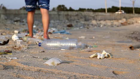 a tripod shot of the legs of a white man who throws a plastic water bottle, which stays on the sandy beach and the man walks away through countless rubbish , thailand