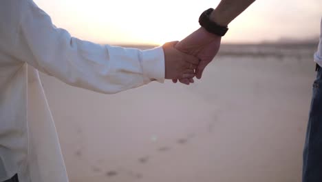 Aimed,-close-up-footage-of-holding-hands-couple-walking-in-romantic-relationship-under-sun-and-blue-sky-in-desert.-Two-young-lovers-walking-by-desert-sand-in-casual-clothes.-Landscape-on-background