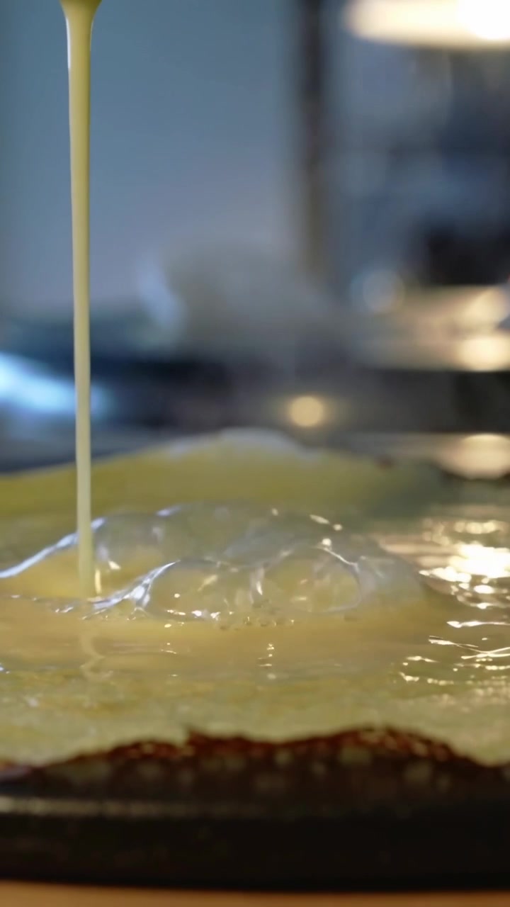 Vertical video: Pouring pale batter via spout making dome on pan, forming lacy brown edge for crepe
