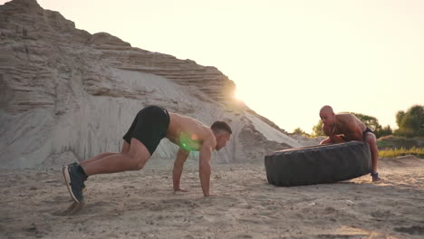 two muscular open-chested athletes train in active mode on the beach doing push-ups and pushing a huge wheel against a sandy mountain at sunset