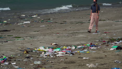 a woman walking on the beach filled with garbage washed up on the sand, static shot