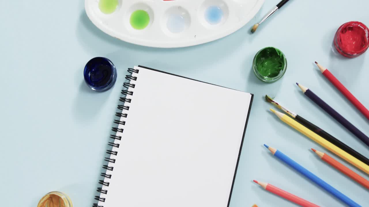 vista aérea del cuaderno, pinceles y pinturas sobre fondo azul, en cámara lenta