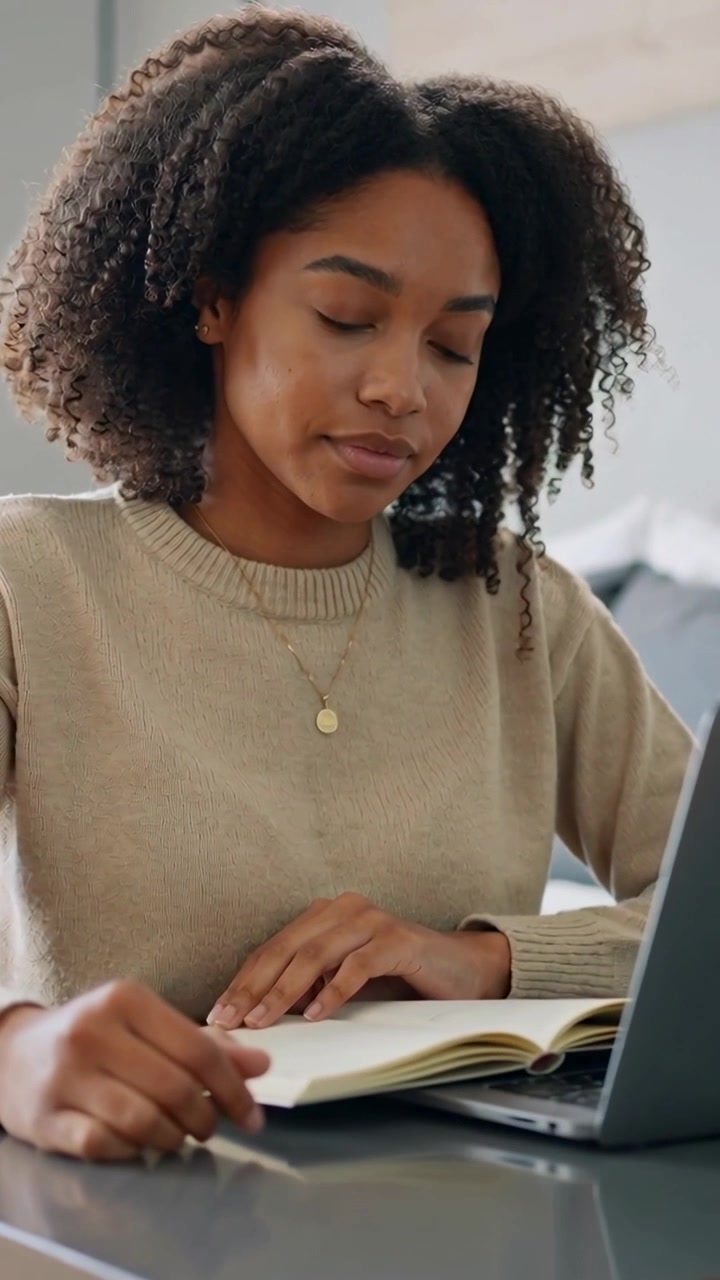 Vídeo vertical: Mulher lendo, de suéter bege, olhando para um livro e um laptop em casa, usando um pingente.