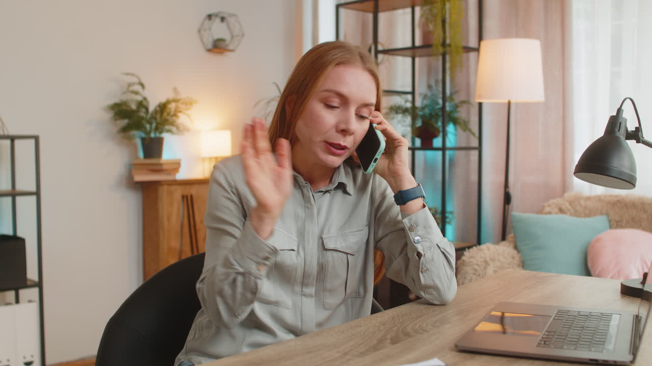Angry caucasian mature businesswoman freelancer talking on smartphone sitting at home office table