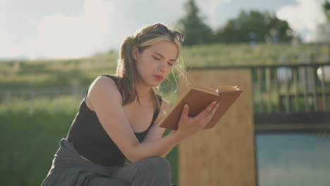 woman seated with legs crossed, reading a book while the wind blows through her hair, her hand rests on the book, and a black bag is beside her, the background features a grassy hill