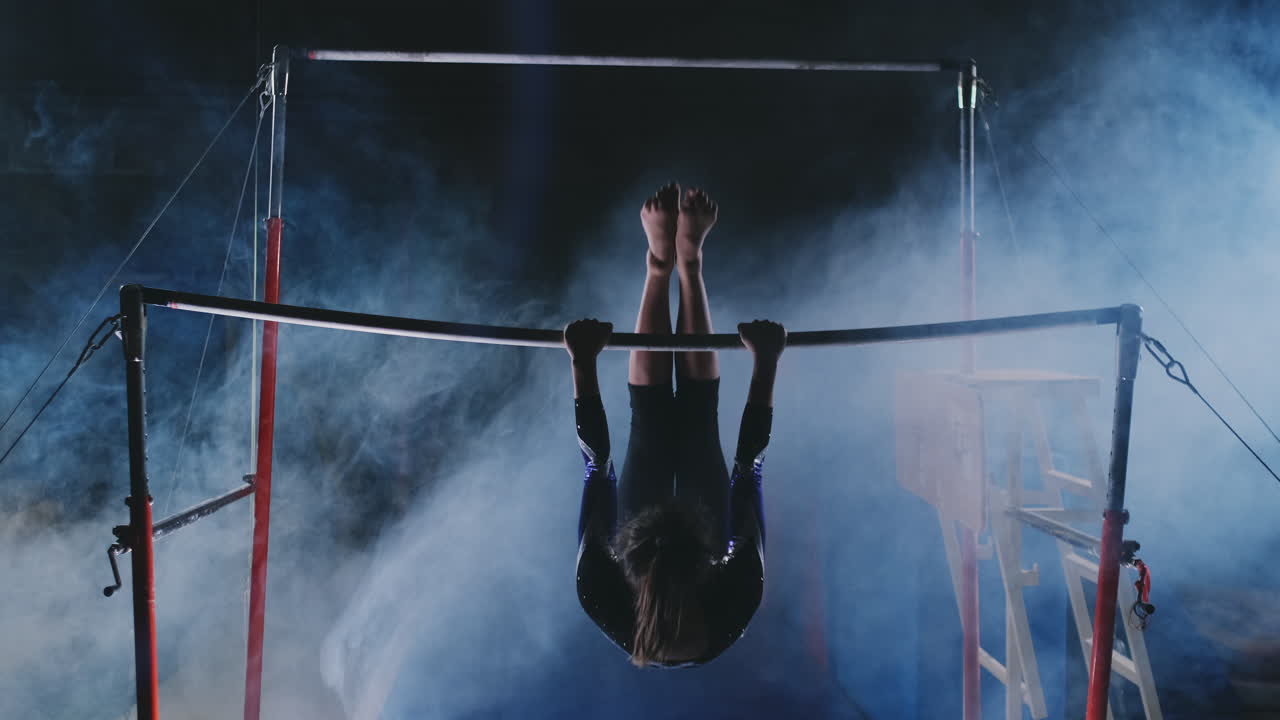 Competizioni di ginnastica. Una donna si avvicina alla sbarra ed esegue flessioni e volteggi al rallentatore su uno sfondo scuro in controluce