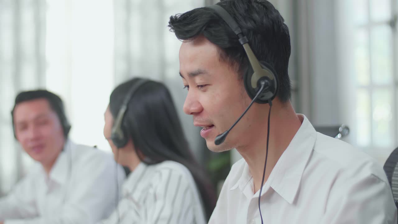 A Man Of Three Asian Call Center Agents Wearing Headset Speaking To Customers On The Call While Two Of His Colleagues Are Talking Working At The Office