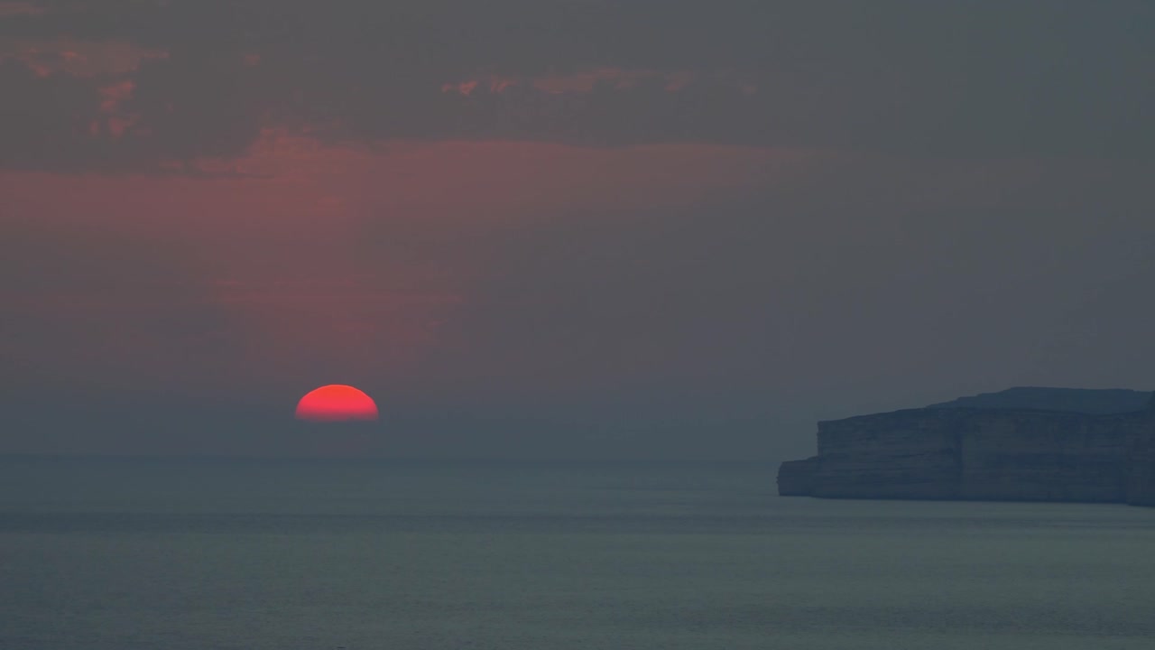 The faintly red setting sun disappears behind the horizon of the Mediterranean Sea off the rocky shores of Gozo on a summer day in Malta.