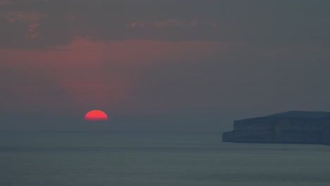 the faintly red setting sun disappears behind the horizon of the mediterranean sea off the rocky shores of gozo on a summer day in malta.