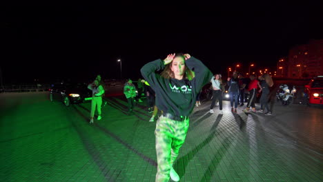group of youngsters dancing in the street at night. beautiful girl moving forward following camera. cars stand at backdrop.