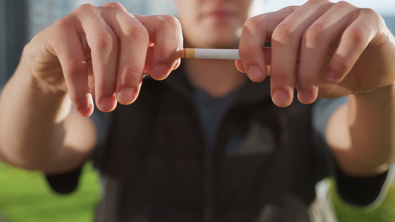 man standing with sunlit hands brought forward gripping and snapping cigarette mid fail gesture in front of blurred building and lush greenery, attempt to quit smoking