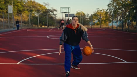 Porträt-Eines-Blonden-Mädchens-In-Sportkleidung-Und-Blauen-Hosen,-Das-Einen-Orangefarbenen-Basketballball-Vom-Boden-Abprallen-Lässt,-Damit-Manövriert-Und-Ihn-Dann-Im-Sommer-Auf-Einem-Roten-Basketballplatz-Auf-Der-Straße-In-Den-Korb-Wirft