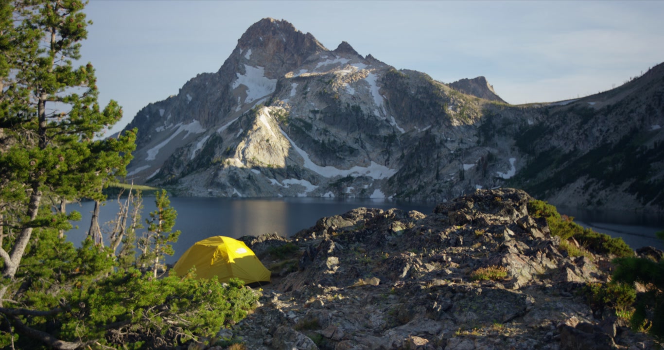 Yellow backpacking camping tent on rocky ridge overlooking Sawtooth Lake and snow capped mountain in Idaho, static nobody