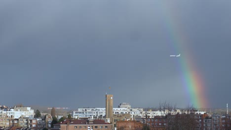 Airplane-flying-through-rainbow-in-the-sky-while-Belgian-flag-waving-at-the-town-hall-of-Woluwe-Saint-Lambert,-Brussels