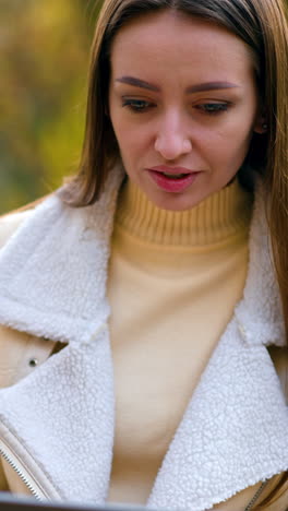modern young woman using laptop sitting in the nature. brunette beautiful lady sitting on the table looking at computer screen. autumn nature backdrop. vertical video