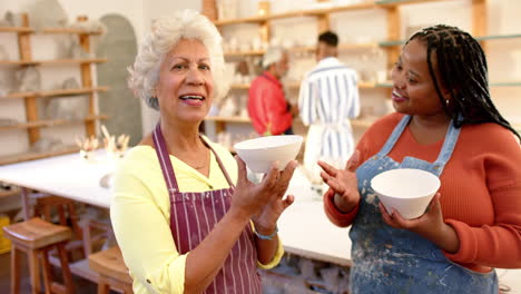 happy diverse female potters holding bowls and smiling in pottery studio, slow motion