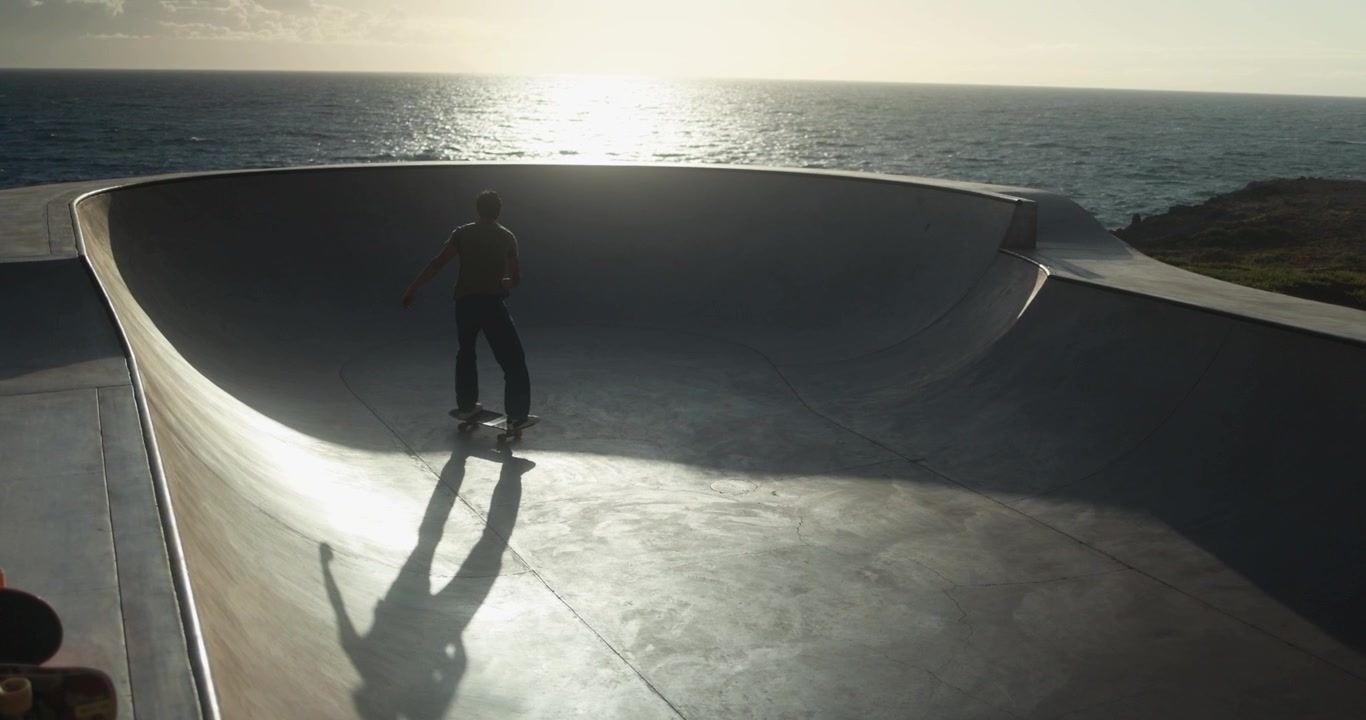 Skateboarder che fa skateboard in una bowl di cemento al tramonto vicino all'oceano
