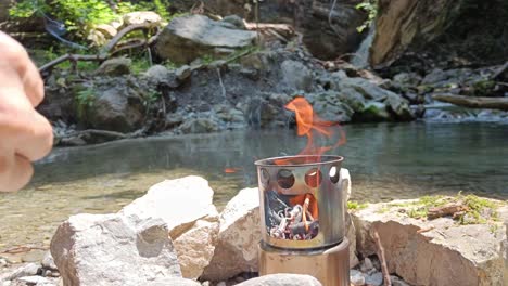 guy adds wood to hobo cooker in nature to cook food, beautiful river and waterfall in background