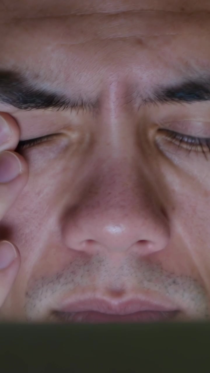 Vídeo vertical: Homem adulto esfregando a mão direita, aliviando a fadiga ocular em um quarto com pouca luz, com o celular brilhando abaixo.
