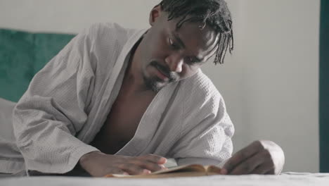 man in white robe smiling while reading large book, lying on bed, enjoying content. soft light filters through window, enhancing warm, peaceful, and relaxed atmosphere in comfortable indoor setting