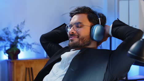 Happy-smiling-Indian-businessman-listening-music-on-headphones-taking-break-relaxing-at-office