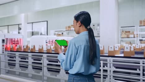 back view of asian female with conveyor for packaging coffee beans in bags at coffee factory, checking on smartphone with green screen and look at the factory