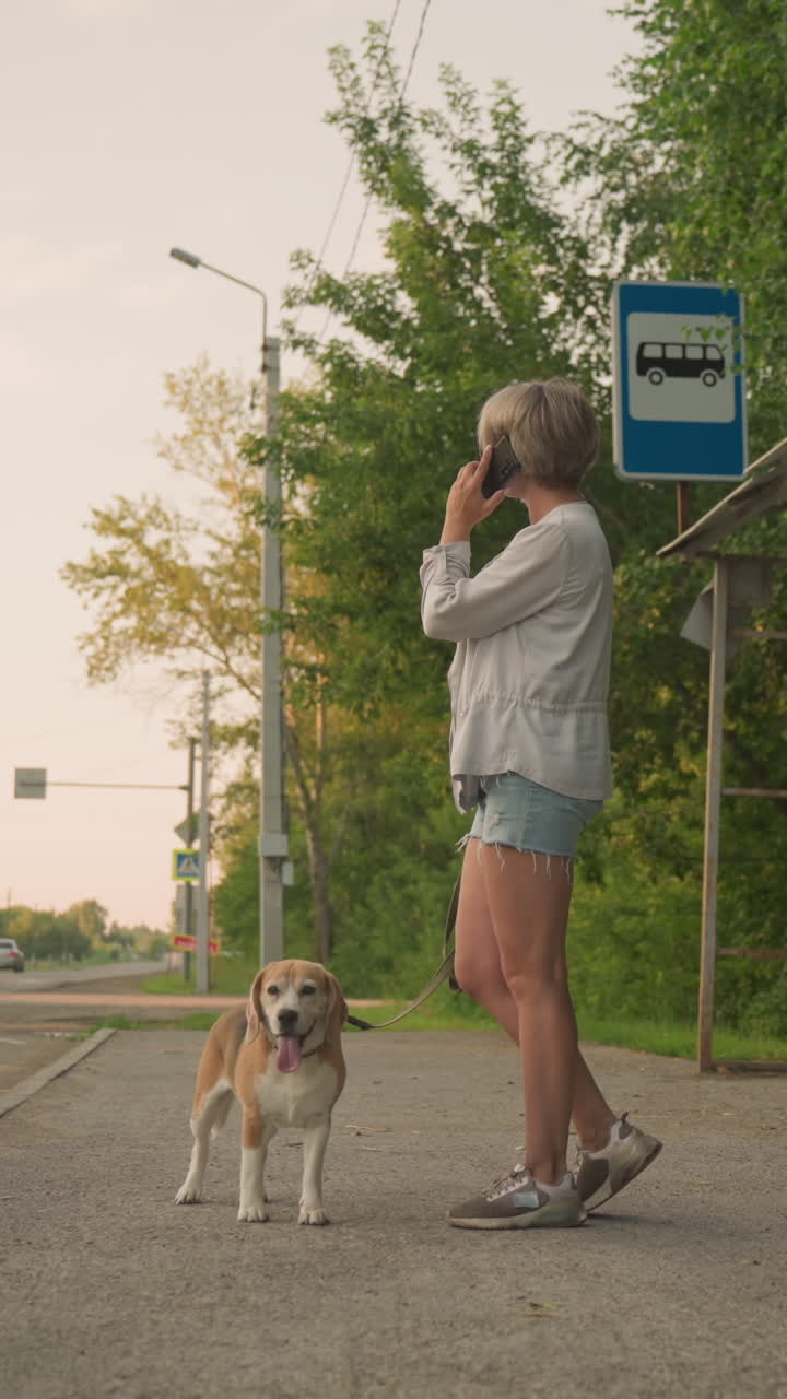 mujer de pie en una parada de autobús en una zona rural sosteniendo a un perro en la correa mientras habla por teléfono, los alrededores incluyen árboles verdes, un refugio de parada de autobuses y una carretera con coches que pasan