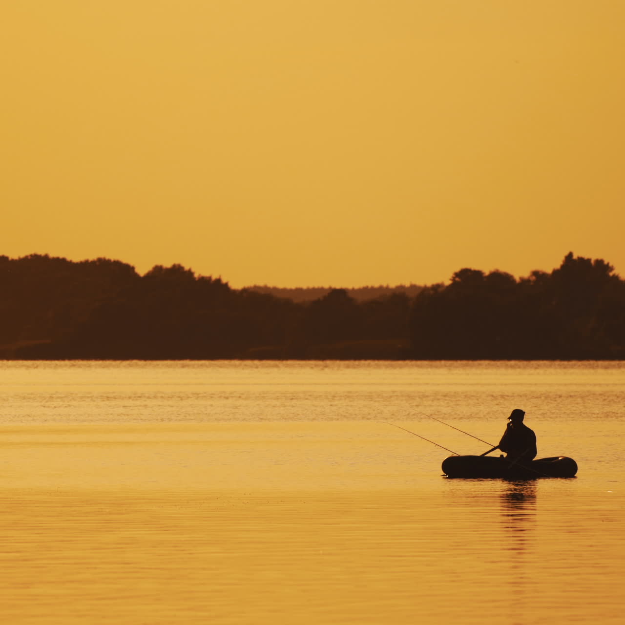 Amazing view of the evening sunset and a man in a boat fishing. Silhouette of a fisherman sitting in a row boat and catching fish on the golden evening background over the river surface.