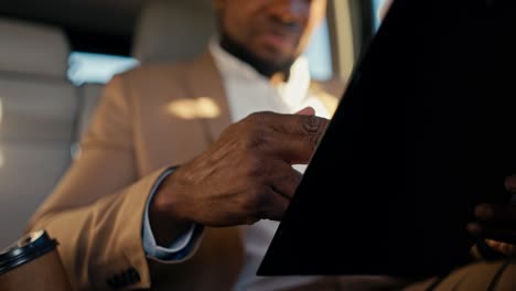 Close-up-of-a-man-Businessman-with-Black-skin-color-enters-a-tablet-with-his-hands-while-presenting-his-plans-while-driving-in-a-modern-car-through-the-city-to-his-destination