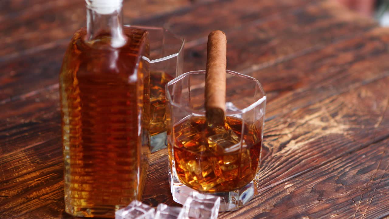 botella de whisky con dos vasos y cigarro cubano colocado sobre una mesa rústica de madera