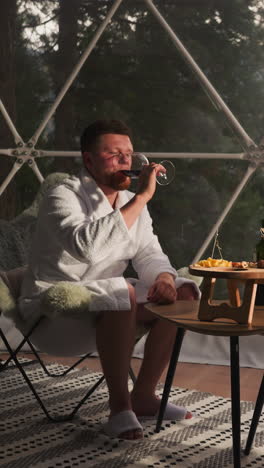 newlyweds drink wine at glamping hotel. couple in terry bathrobes celebrate anniversary on glass terrace. stunning view adds romantic touch to date