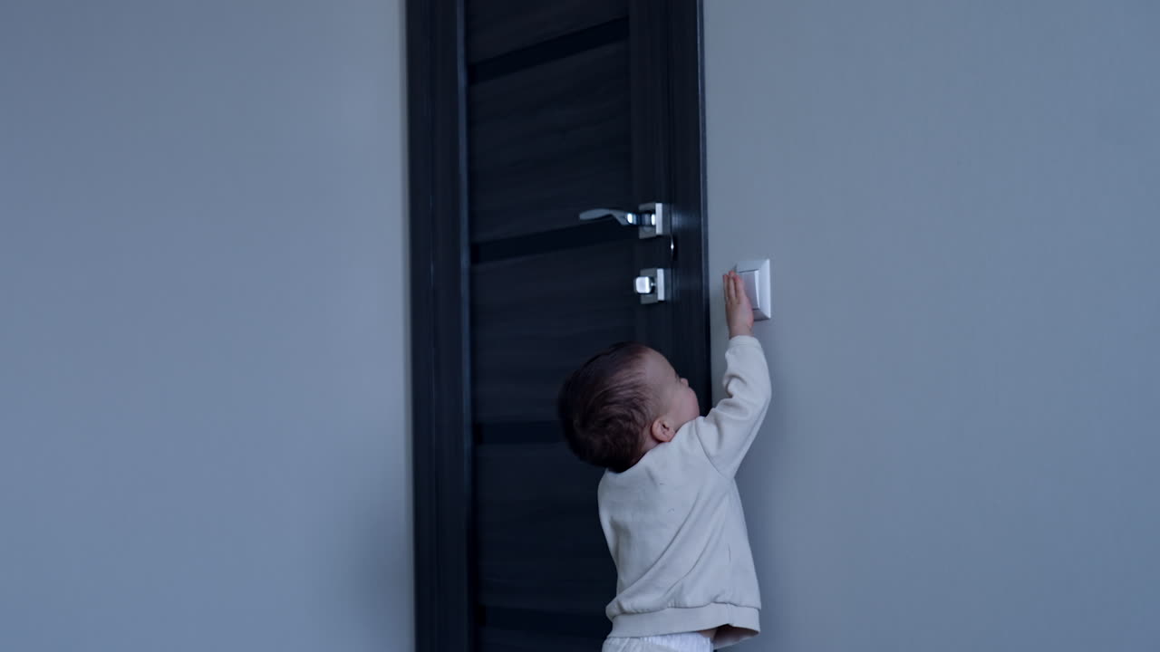 Toddler boy walks up to a light switch. Baby switches the light on cheering and smiling happily.