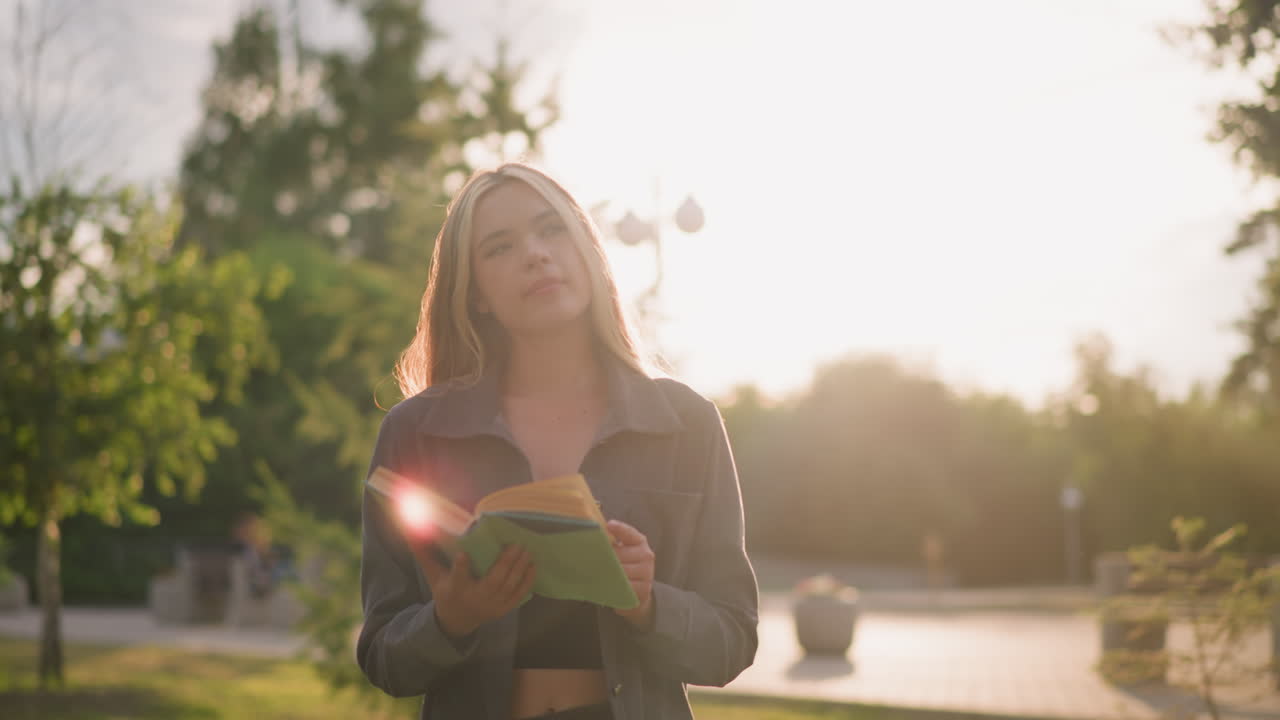 dama con ropa gris caminando por un campo de hierba mientras lee un libro mientras la luz del sol crea destellos brillantes a su alrededor, mirando pensativa, con fondo iluminado por el sol y árboles borrosos por la luz natural