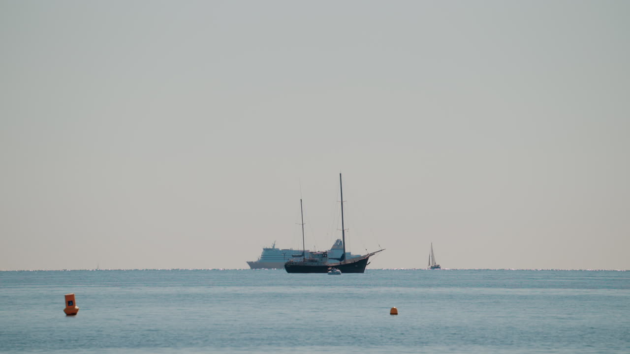 Wide shot of a calm sea with a sailboat and a large cruise ship sailing in the distance under a pale blue sky