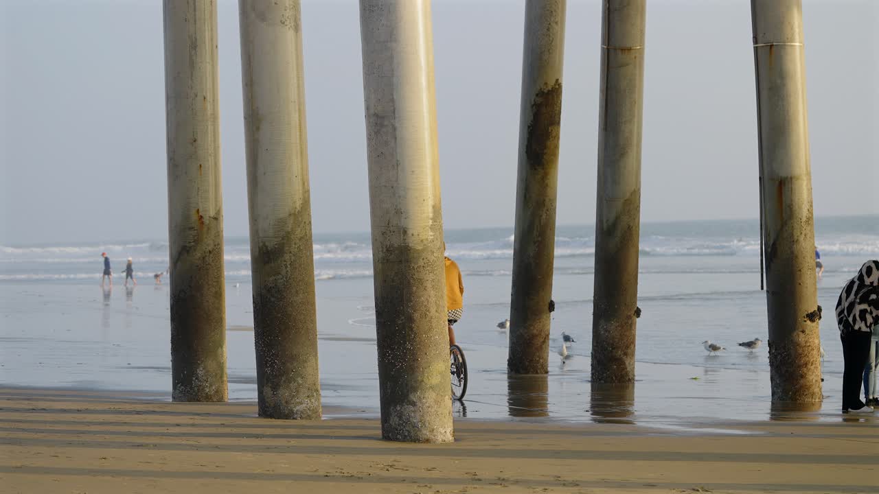 hombre caucásico alto y flaco montando en bicicleta, en la playa, debajo del muelle