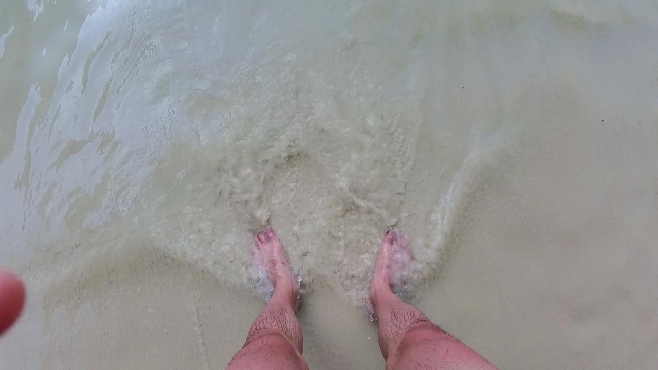 Synspunkt af fødder, der står i blødt hvidt sand med blidt havvand, der skyller ind på land ved Ko Hong Island, Thailand.