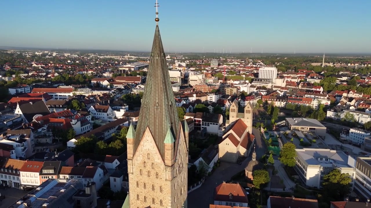 Smooth drone flyby passes the cathedral facade as golden morning sunlight illuminates the warm sandstone and Paderborn skyline behind it.