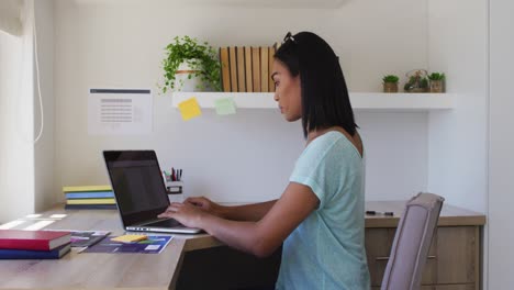 Mixed-race-gender-fluid-person-sitting-at-desk-working-from-home-using-a-laptop