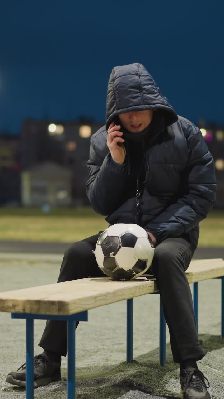 A man is sitting alone on a bench in an empty stadium at night, with a soccer ball between his legs, he reaches into his jacket to retrieve his phone to answer a call