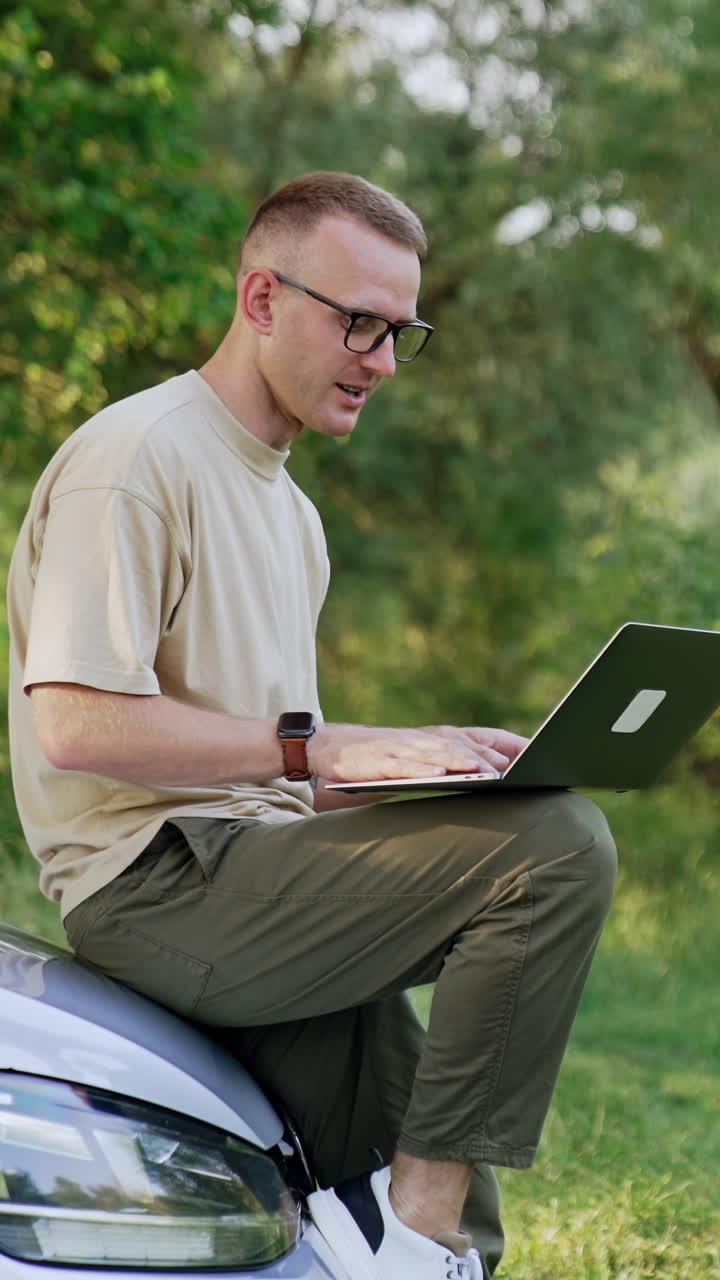 Caucasian male sitting on the car front holding a laptop. Freelancing man types on computer and smiles. Vertical video