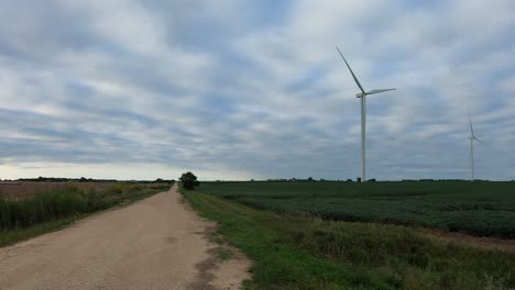 Eine-Sich-Drehende-Windkraftanlage,-Die-Sich-An-Einem-Bewölkten-Tag-In-Einem-Sojabohnenfeld-Entlang-Einer-Schotterstraße-Im-Ländlichen-Nebraska,-USA,-Befindet