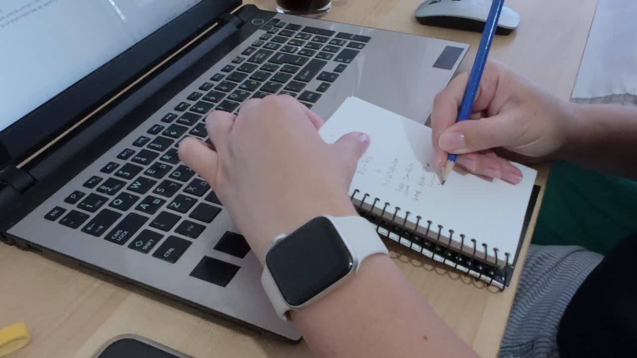 Close-up of hands multitasking, writing in notebook while using laptop