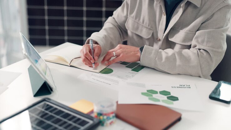 Person Writing in Notebook While Analyzing Business Documents on Desk