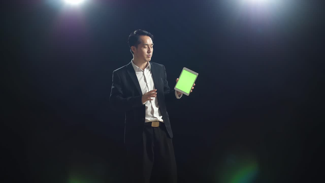 orador asiático en traje de negocios sosteniendo y señalando una tableta de pantalla verde mientras habla en el estudio de pantalla negra
