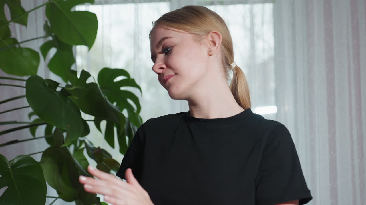 Close up of massage therapist in black shirt , standing calmly in front of large indoor plant near sheer window curtain, expressing peaceful focus and natural wellness environment