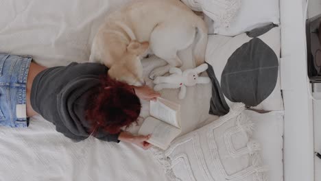Woman relaxing at home with dog, bonding with pet. Owner kissing dog, dog showing affection by licking hands. Reading a book in bed, cozy pet-friendly home, human-animal connection, relaxing moments.