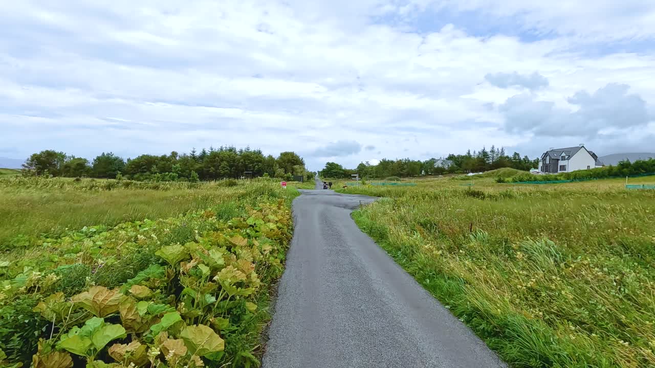 Two motorcyclists travel along a narrow, winding rural road through green fields under cloudy daylight, captured in a steady, wide-angle perspective